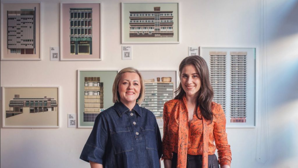 Rachel Loughran and Natalie Tweedie stand in front of Natalie Tweedie's illustrations for the Brutalist Glasgow exhibition at the Glasgow Heritage Trust