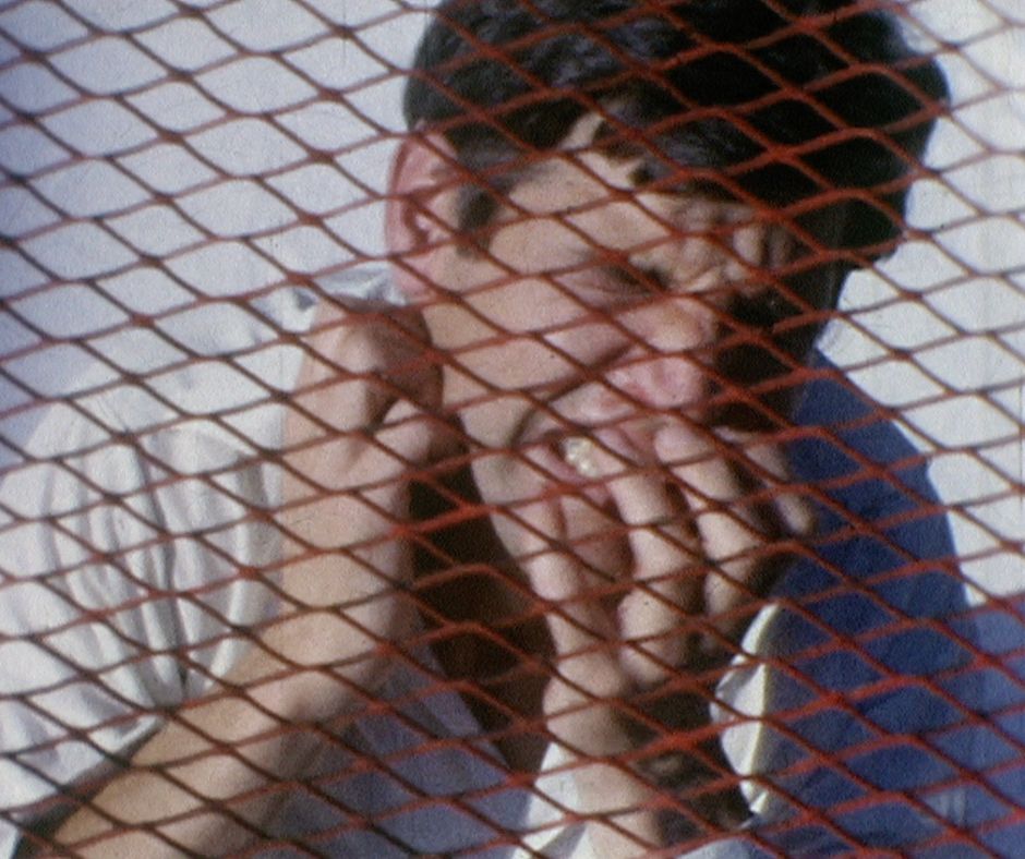 A man with dark hair wearing a white shirt stands behind a red chain link fence, with his eyes tightly shut.