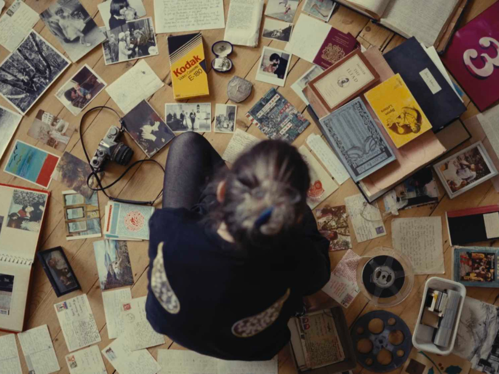 A birds eye view of a woman with her hair in a bun, sitting on the floor with crossed legs, surrounded by old letters and photographs.