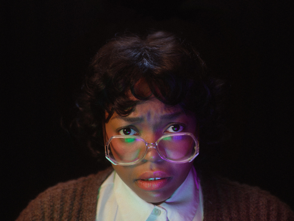 A close-up film still of a woman with dark hair and glasses wearing office attire. Her face is illuminated in purple light and the background is dark.