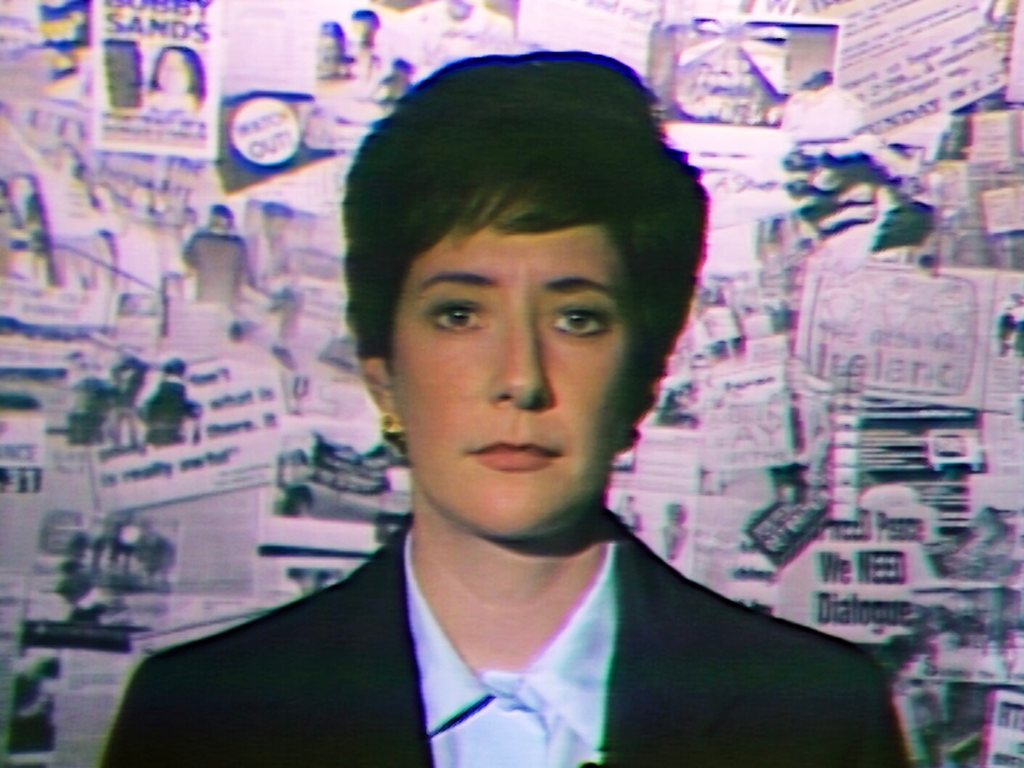 A close-up film still from Frank Sweeney's 'Few Can See'. A woman with short dark hair, wearing a blazer, looks to the camera against the backdrop of newspaper clippings. 