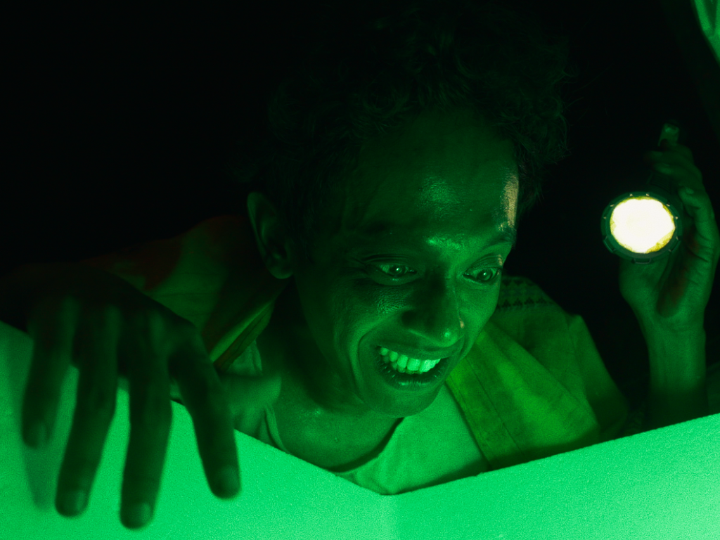 A close-up film still. A man peers into a box, looking excited. Its contents illuminate his face in green light. 